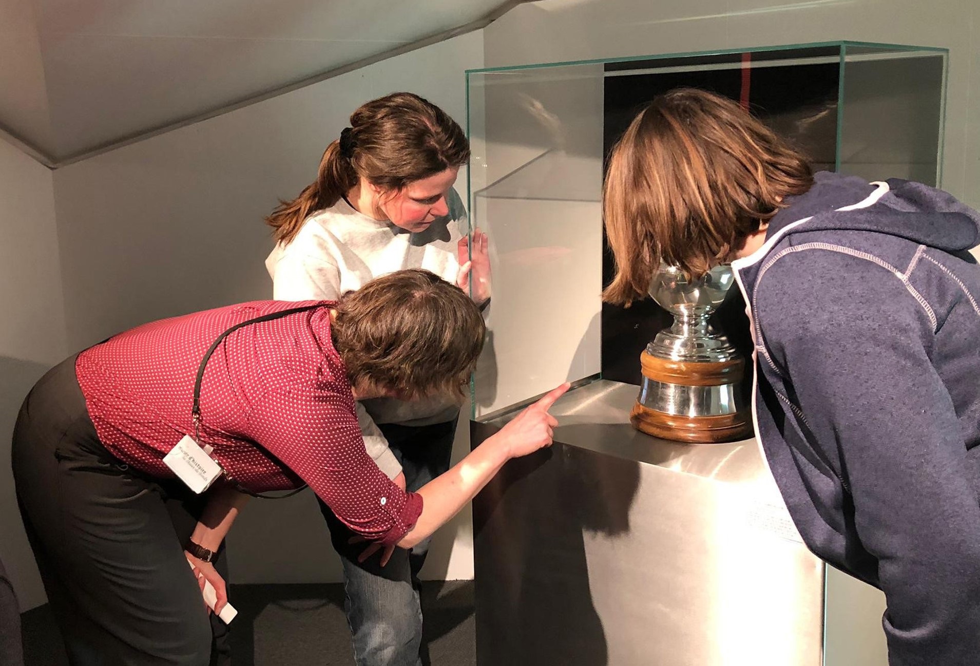 Un objet de la taille et de la forme d’un trophée est exposé à mi-hauteur dans une vitrine. Trois personnes sont penchées vers l’objet, le cachant partiellement. L’une d’elles montre l’objet du doigt.