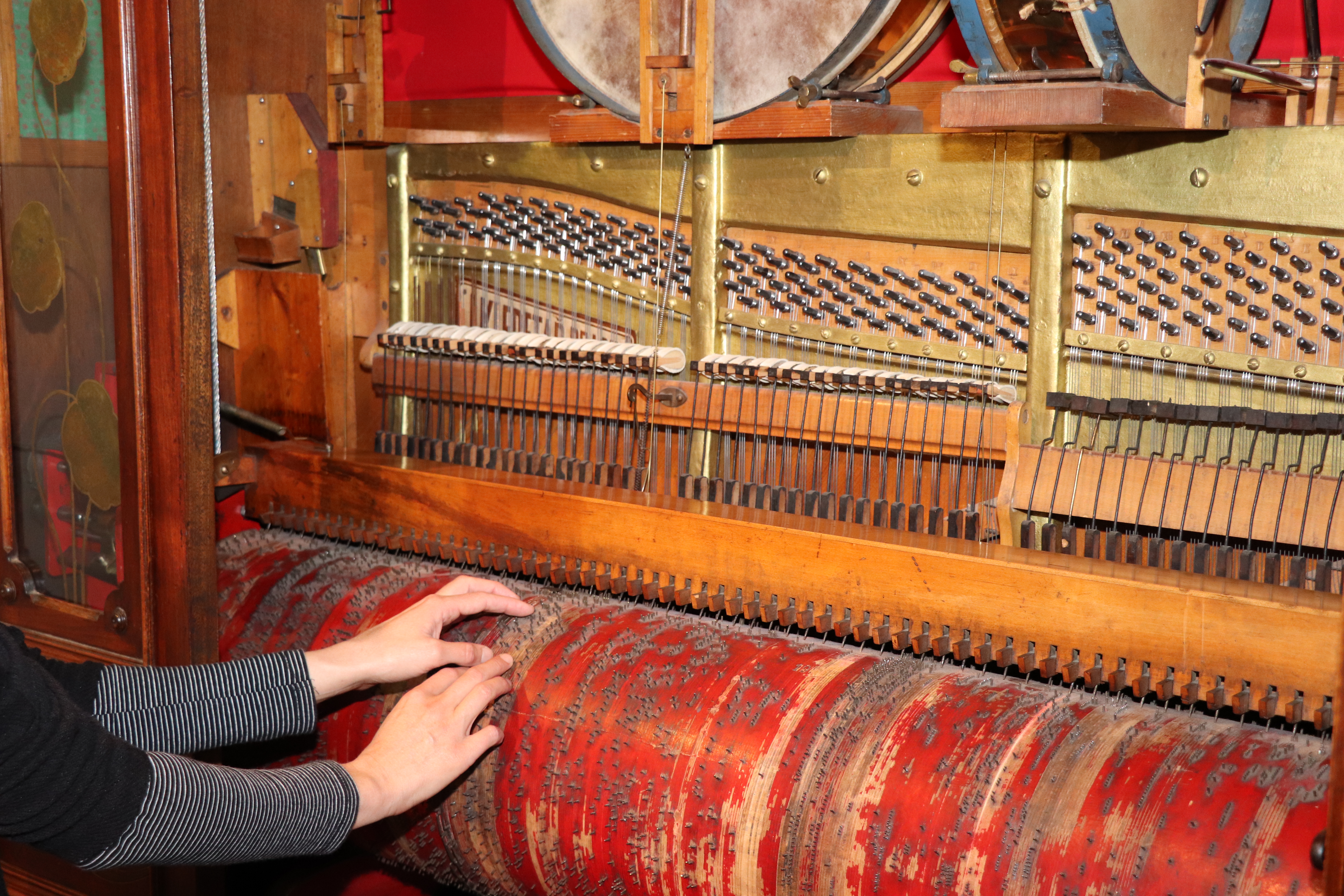 Innenleben eines Musikautomaten: Unten zwei Hände, die eine rote Walze berühren, darüber eine Holzverklei-dung, welche horizontale Saiten und eine darüberliegende vertikale Reihe Knöpfe einrahmt. Eine goldfarbene Metallverkleidung umrahmt die Knöpfe.