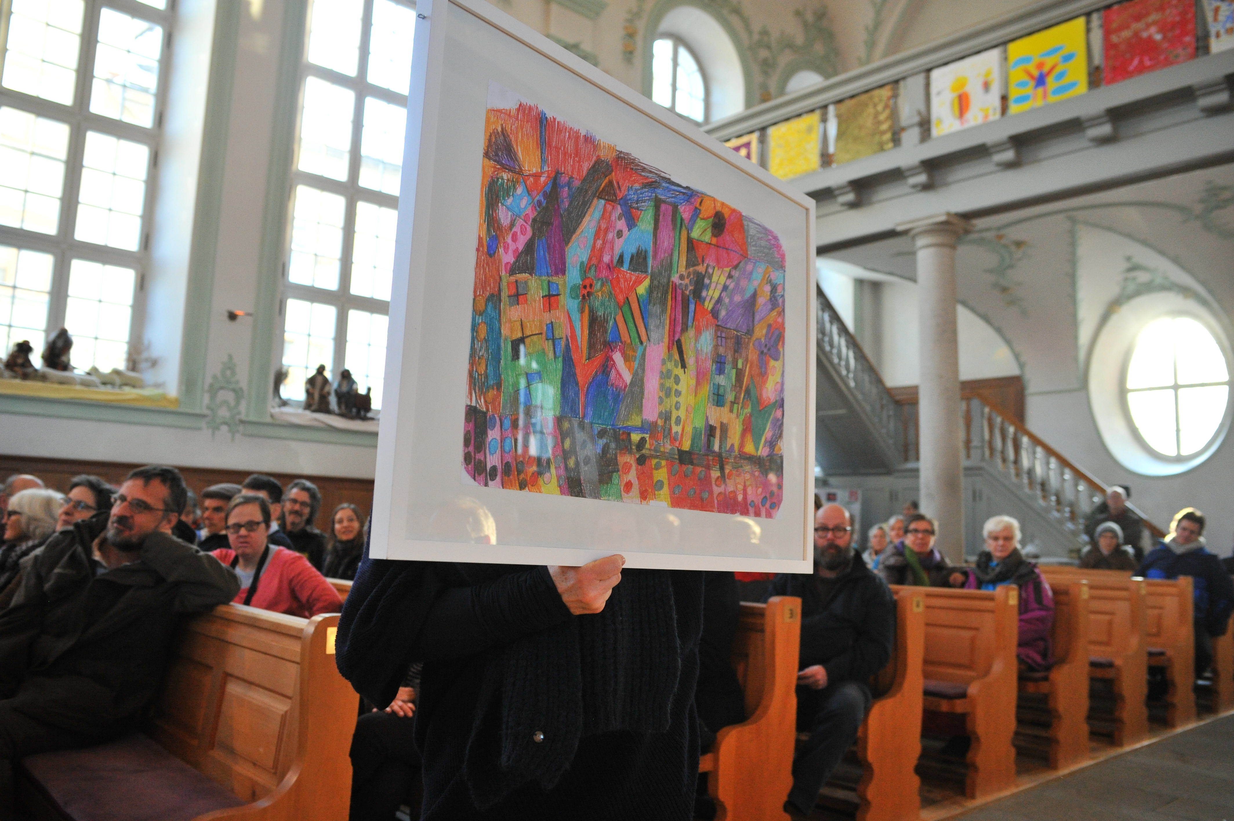 Blick auf ein Kirchenschiff mit zahlreichen Menschen auf den Bänken. In der Bildmitte hält eine Person ein mit Buntstiften angefertigtes, abstraktes farbenfroh gestaltetes Bild hoch, das weiss gerahmt ist. An der Empore sind weitere Bilder aufgereiht.