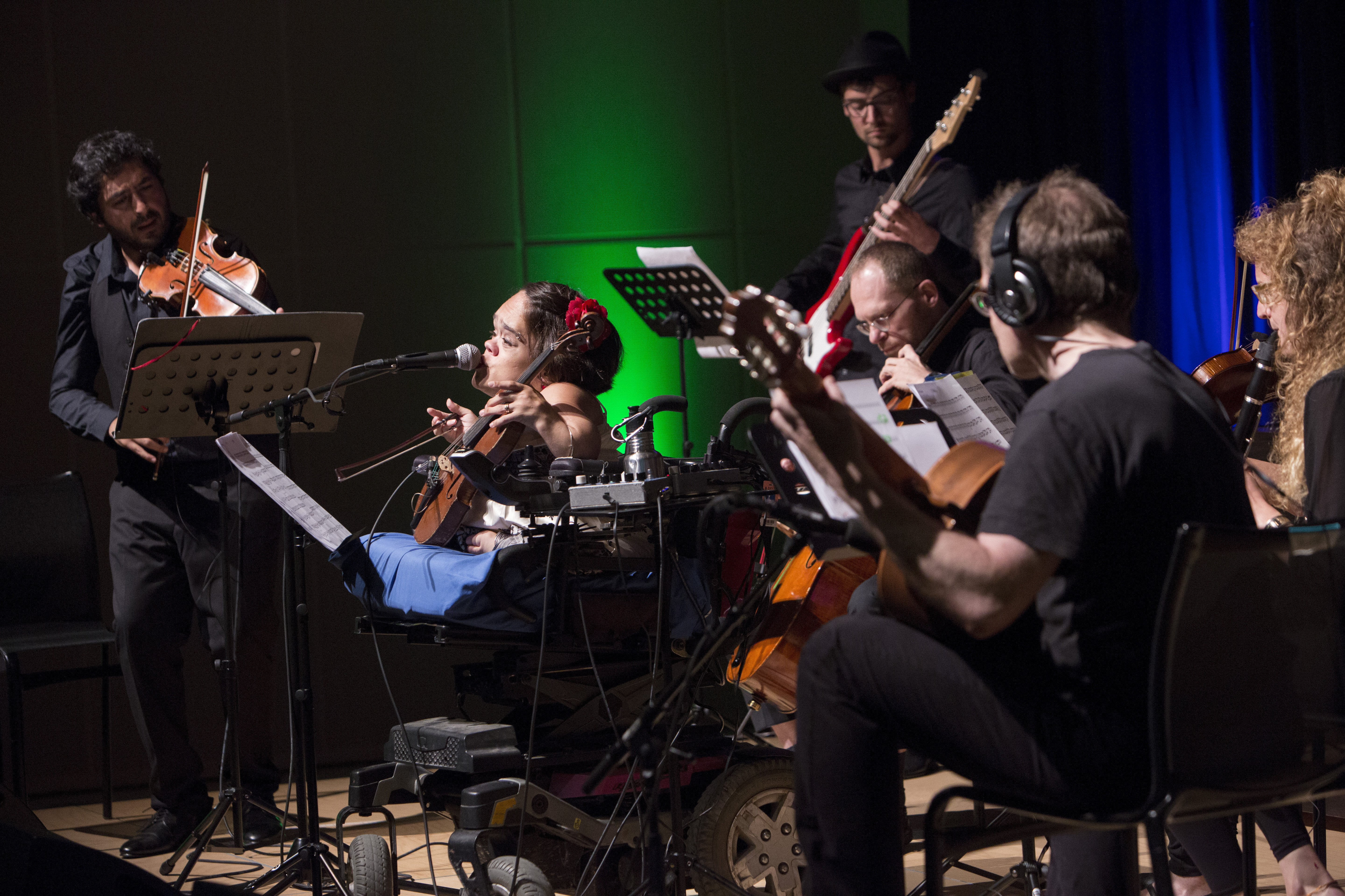 Sechs Personen spielen auf Streichinstrumenten, klassische und E-Gitarre, Klarinette. Fünf in Schwarz, die Frau im Zentrum trägt Weiss, eine rote Blume im Haar. Ein Mikrofon nah am Mund, die Geige stützt sie auf dem Sitz des Rollstuhls. Sie ist klein.