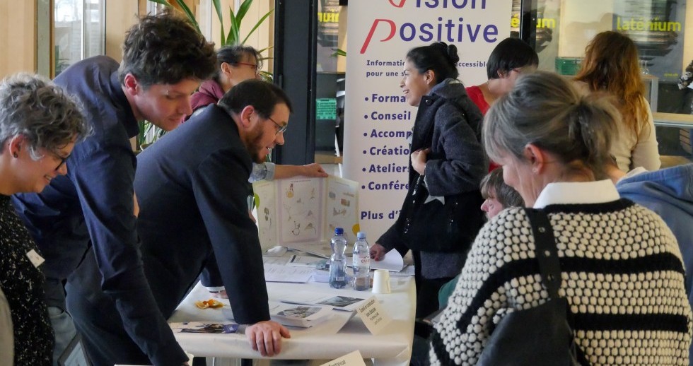 Un hall avec des baies vitrées et de la lumière naturelle. Devant et derrière des tables, des personnes discutent par groupes de deux. D’autres personnes regardent avec intérêt des dépliants sur les tables. 